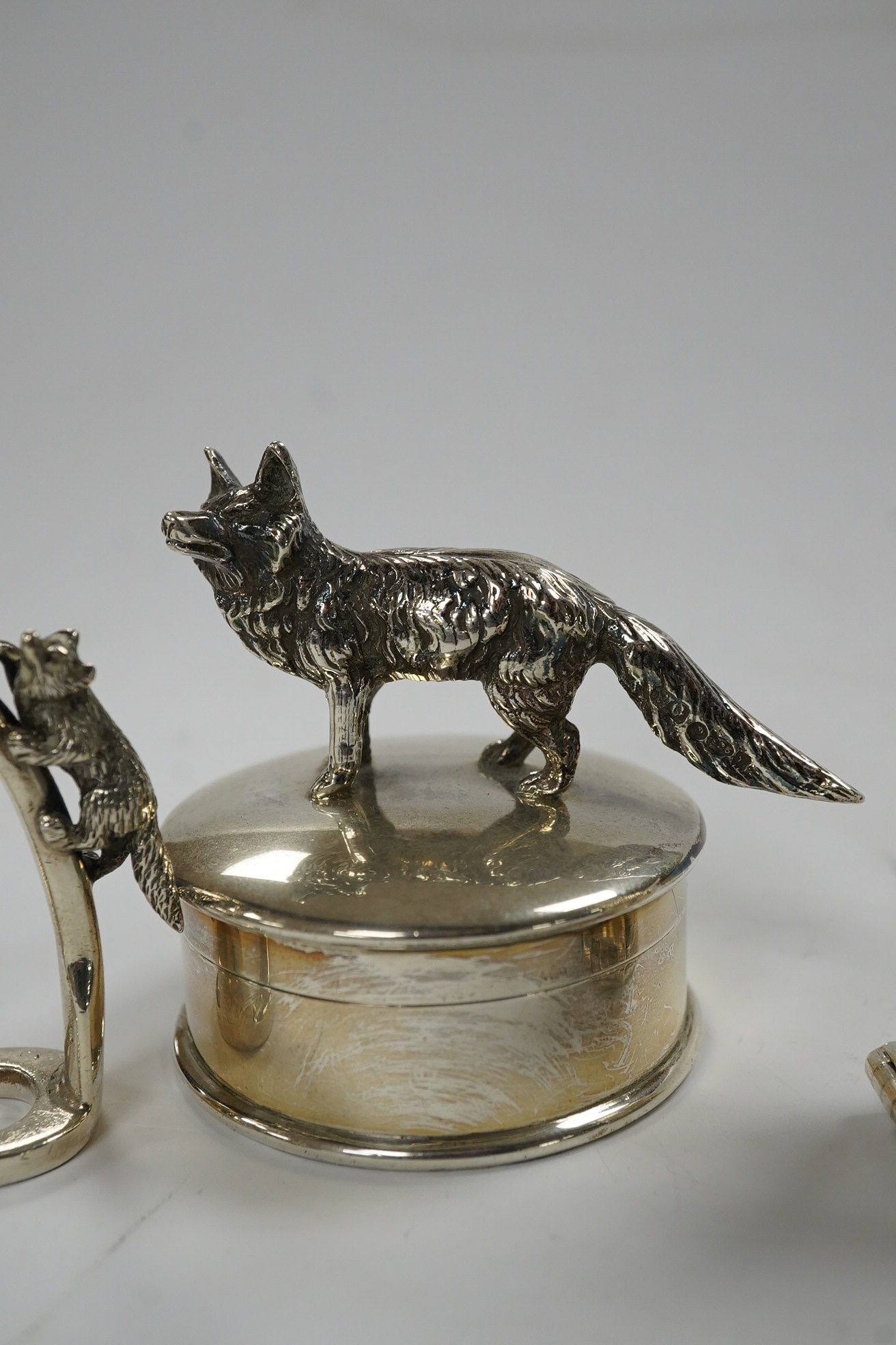 A George VI silver napkin ring modelled as a stirrup, surmounted with a fox, Birmingham, 1937, 46mm, together with a later silver horses head snuff box and a silver pill box with fox surmount. Condition - fair to good